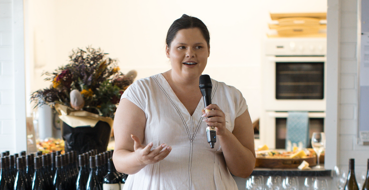 Sommelier Nina Throsby speaking into a microphone in front of rows of wine bottles