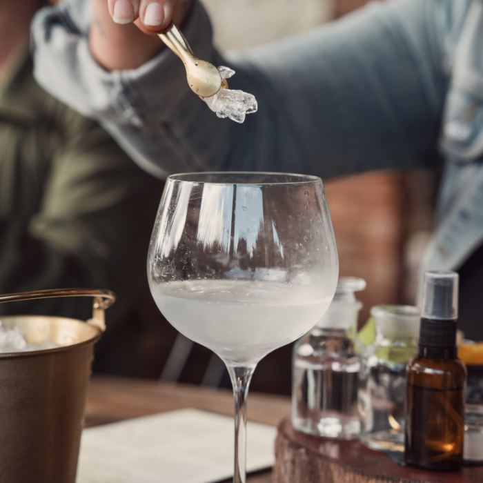 Person adding ice to a wine glass with various bottles and items on a table.