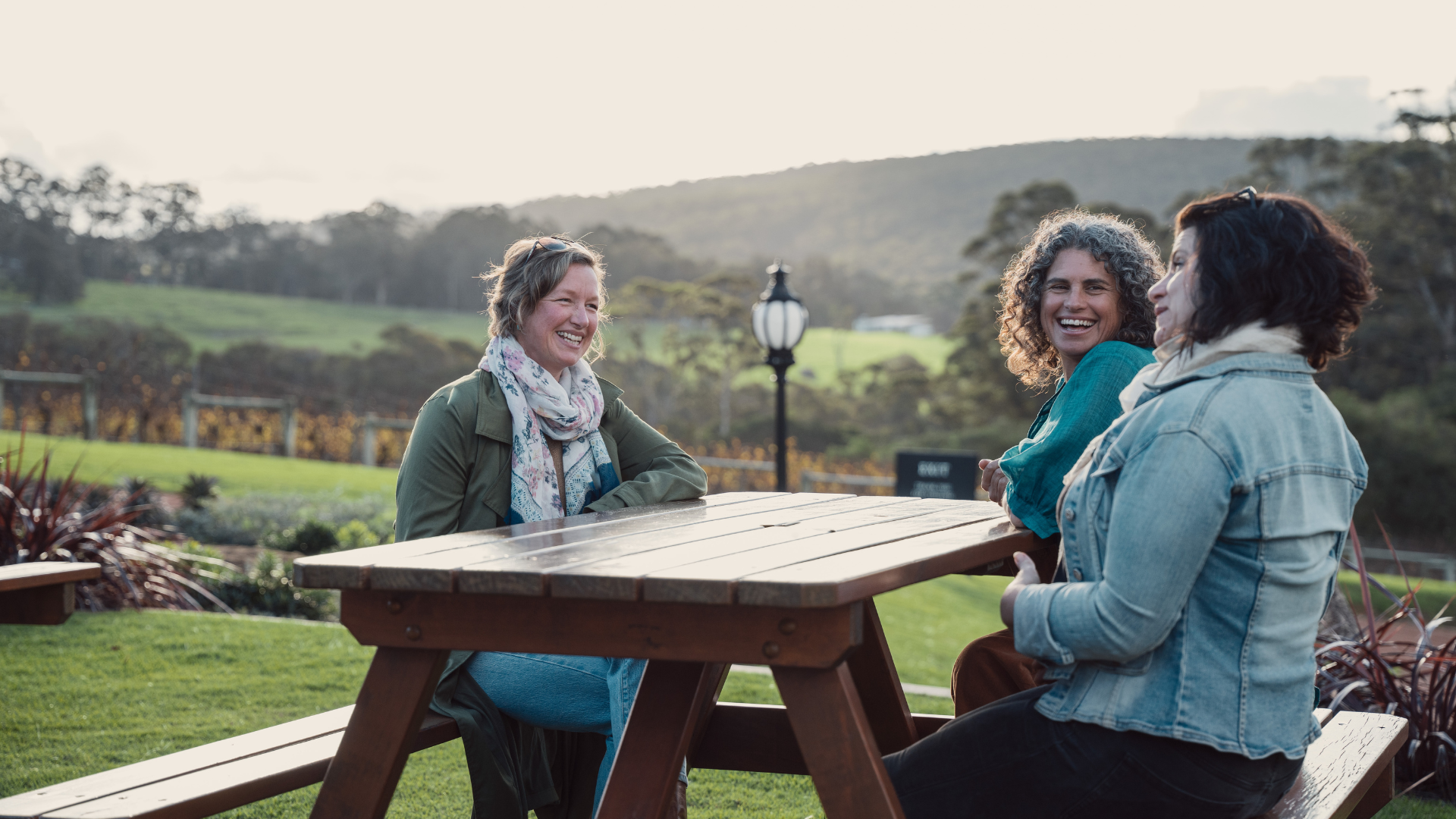 Three women sitting at a wooden table outdoors with a scenic background