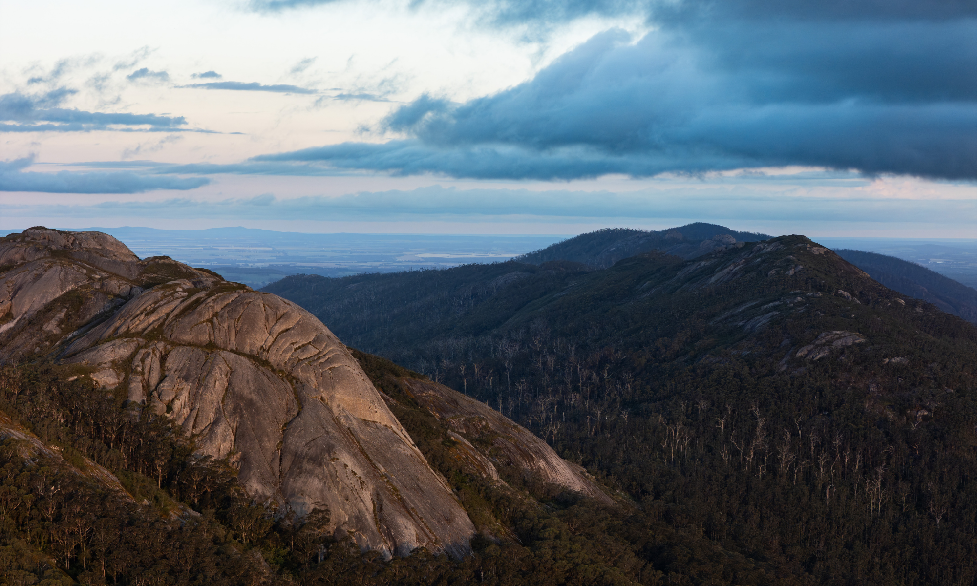 Mountain landscape with a large rock formation and distant mountains under a cloudy sky.