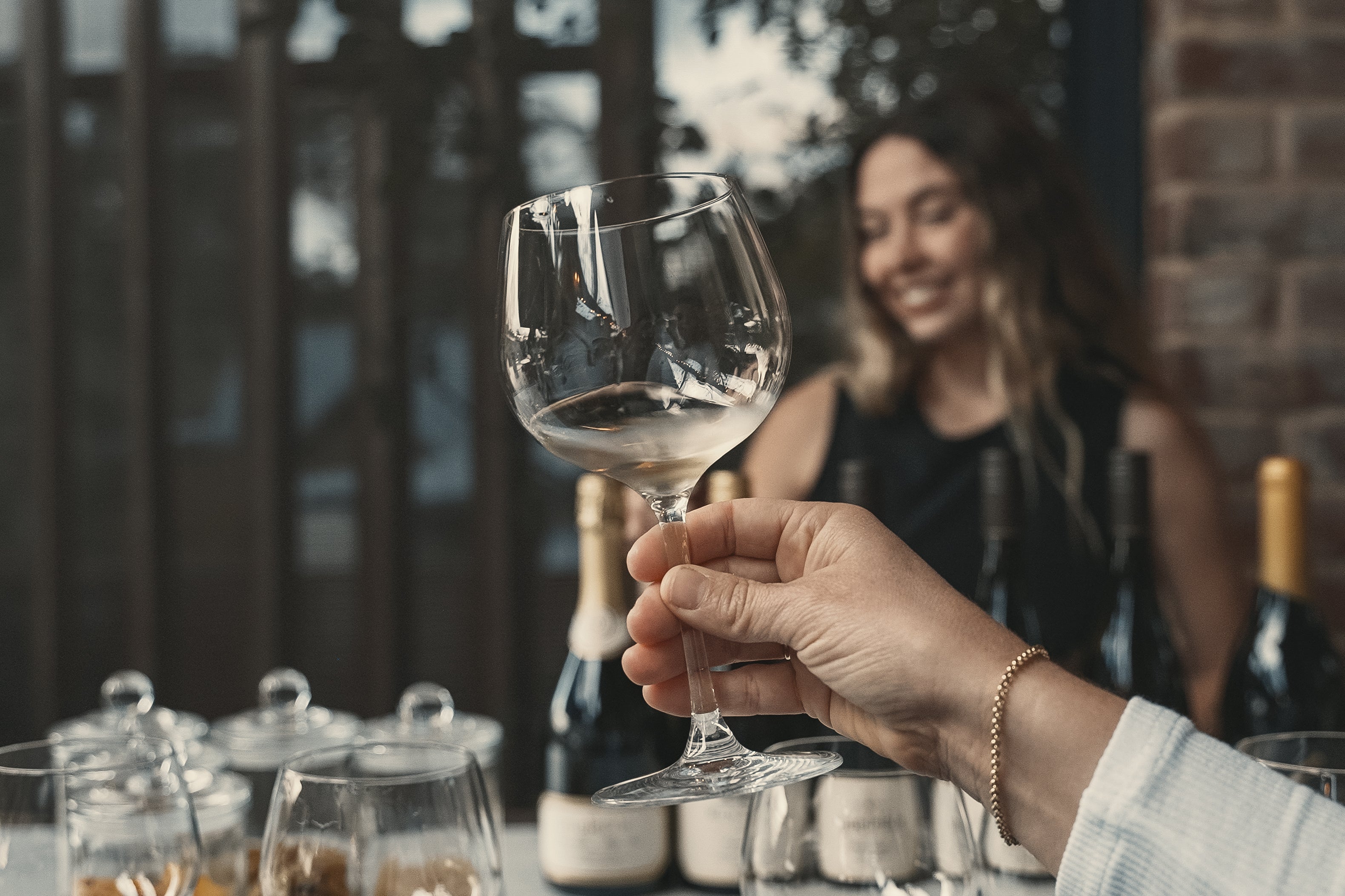 Hand holding a wine glass with blurred background of a woman and wine bottles.