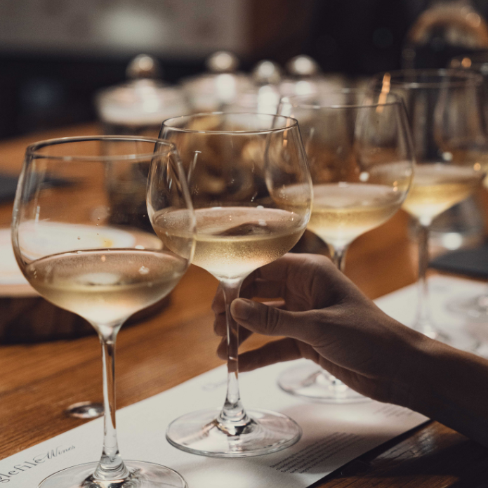 Hand holding a wine glass with white wine on a wooden table, surrounded by other glasses.