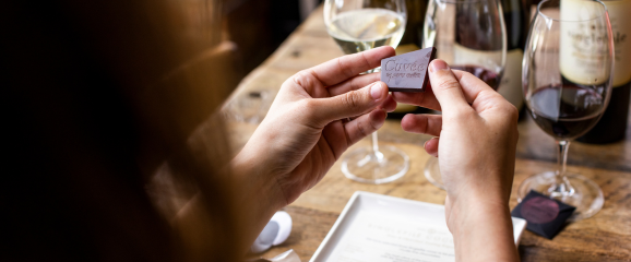 Person holding a wine glass label with wine glasses and bottles in the background