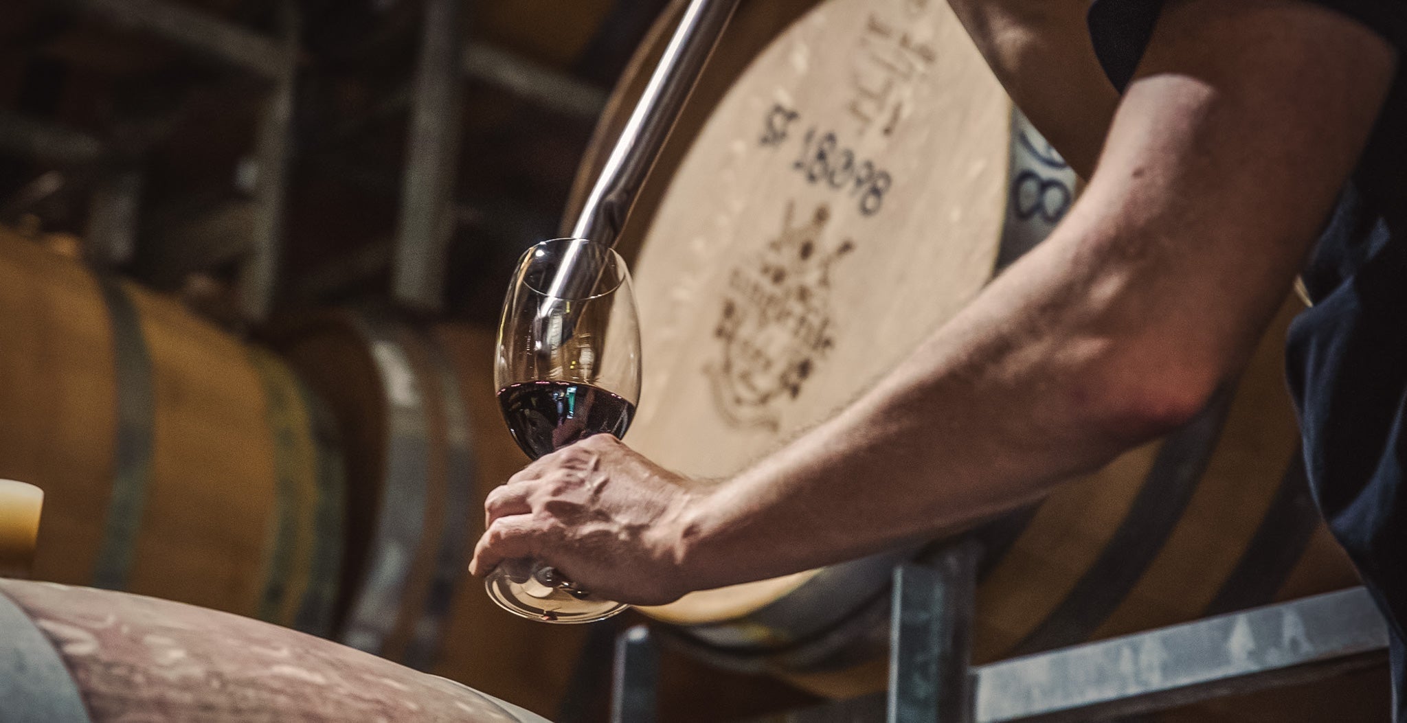 A stainless steel wine thief released red wine in to a glass in front of a wine barrel