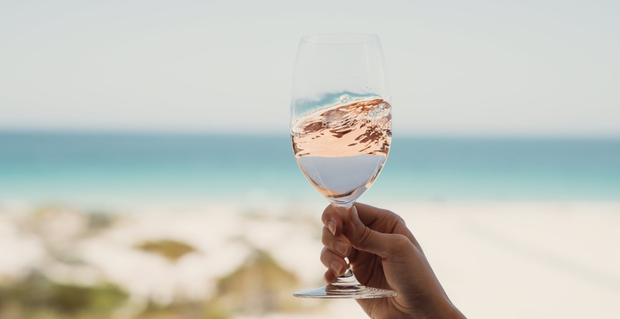 A hand swirling a glass of Rosé with the ocean in the background