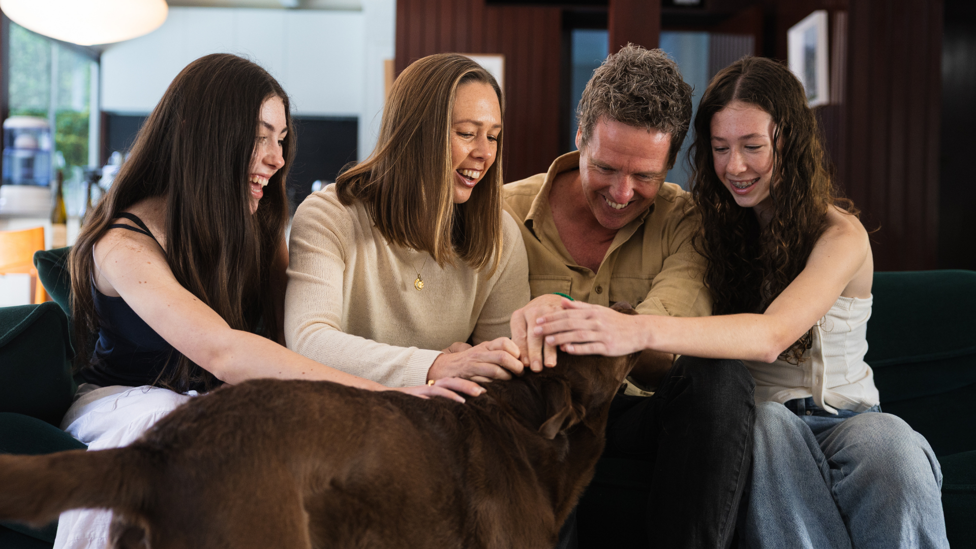 Four people sitting on a couch with a dog, smiling and petting it.