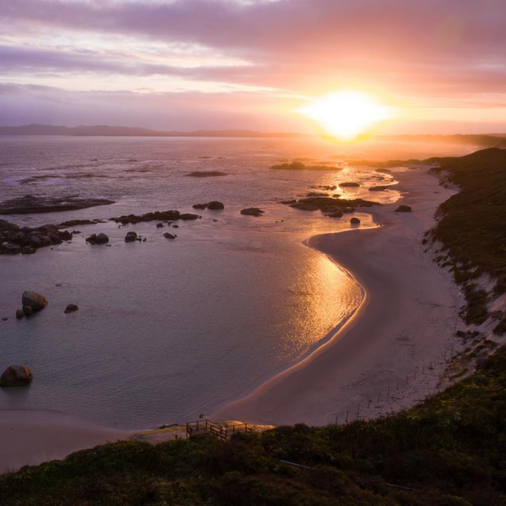 Sunset over a beach with rocks and a curved path leading to the water.