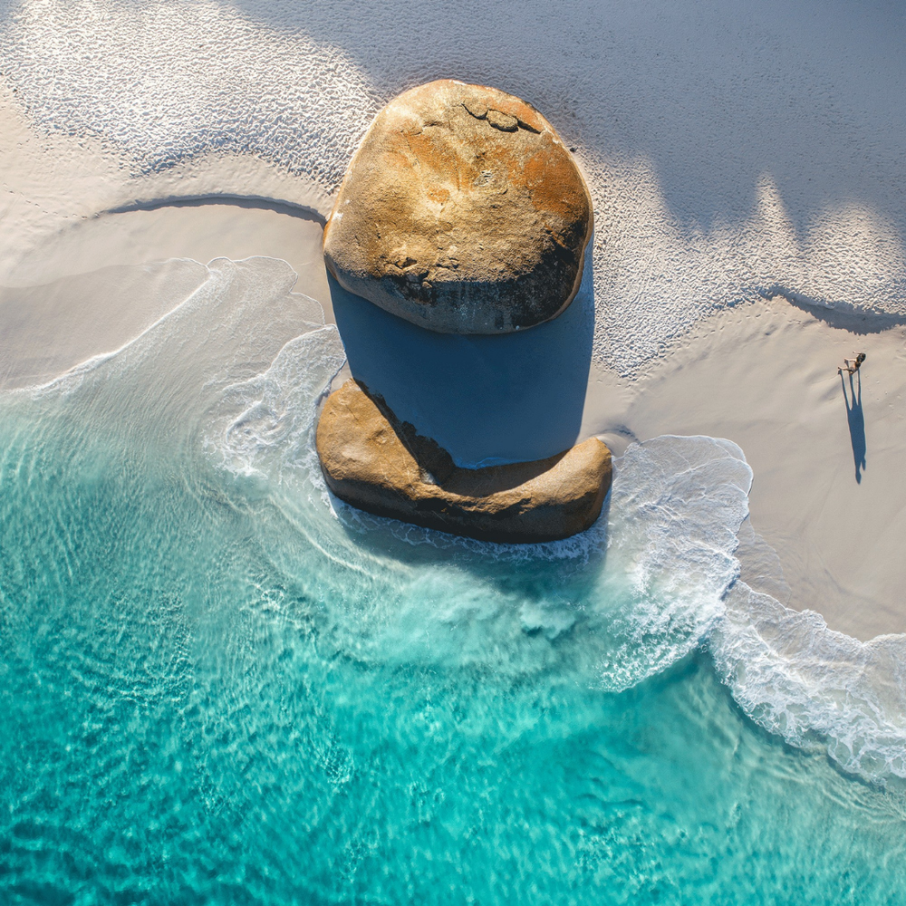 Aerial view of a large rock in clear blue water with a person standing nearby on a sandy beach.