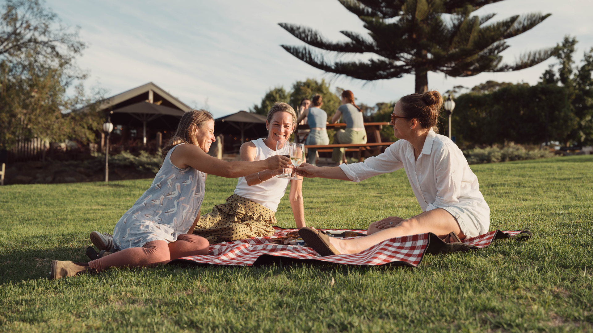 Three people enjoying a picnic on a grassy field with trees and a building in the background.