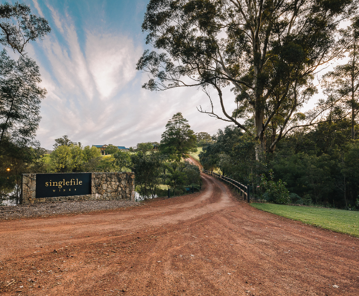 Winding dirt road leading to a stone sign with 'singlefile wines' in a scenic area.