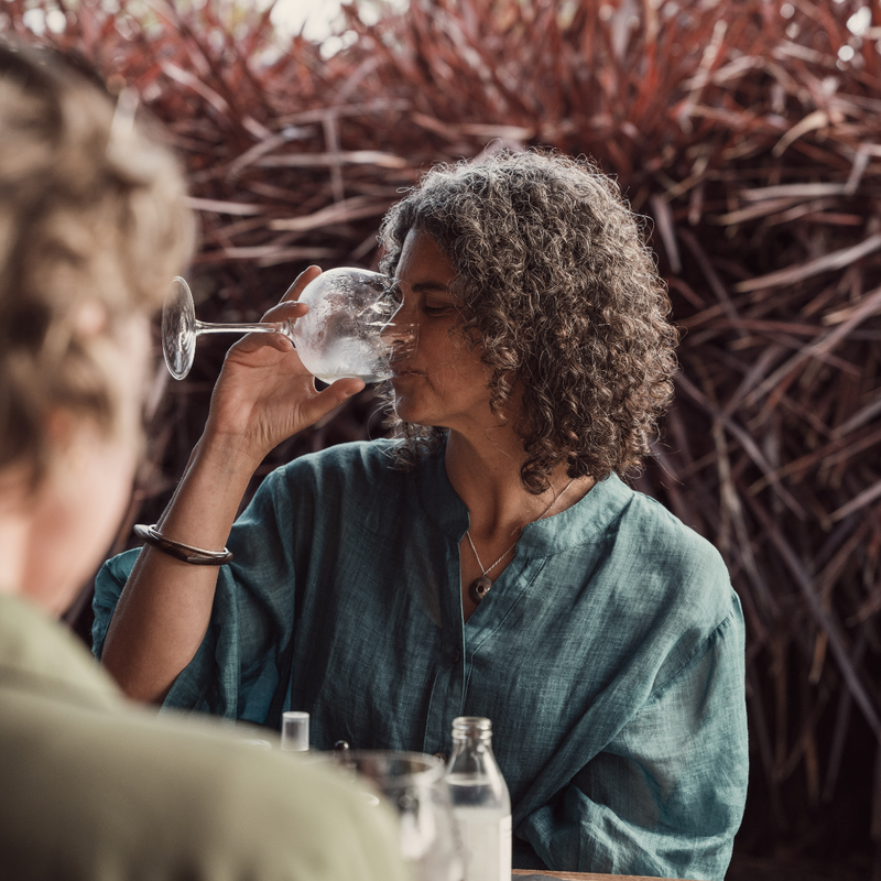 Woman drinking from a glass with another person partially visible in the foreground, against a natural background.
