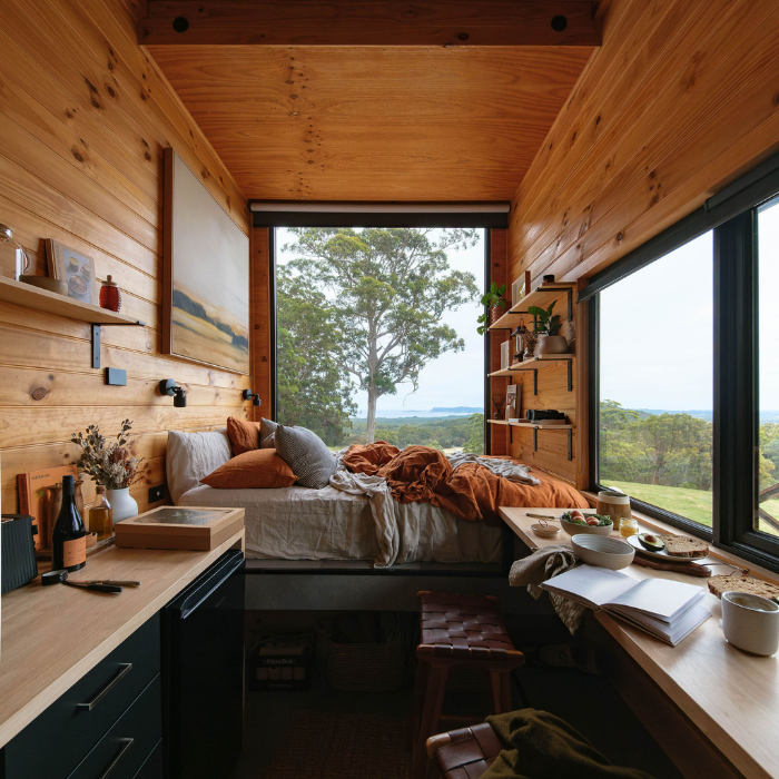 Wooden cabin interior with a bed, desk, and large windows overlooking nature.