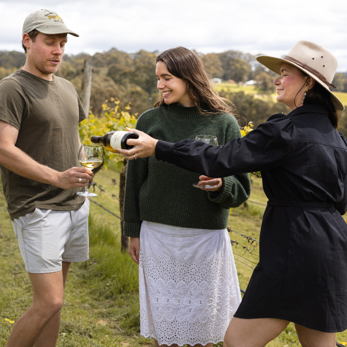 Two women and a man in a vineyard setting, with one woman pouring wine.